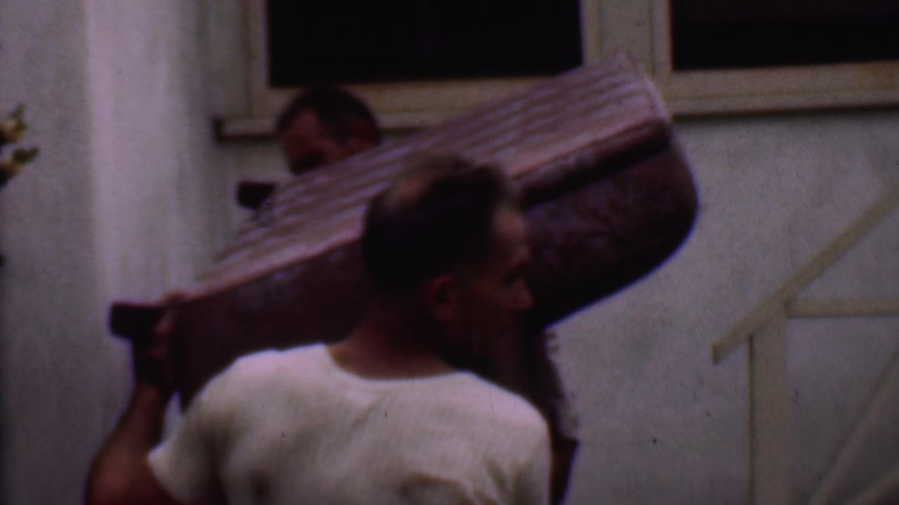 A man in a white shirt supports a large couch while another man is partly visible behind it.