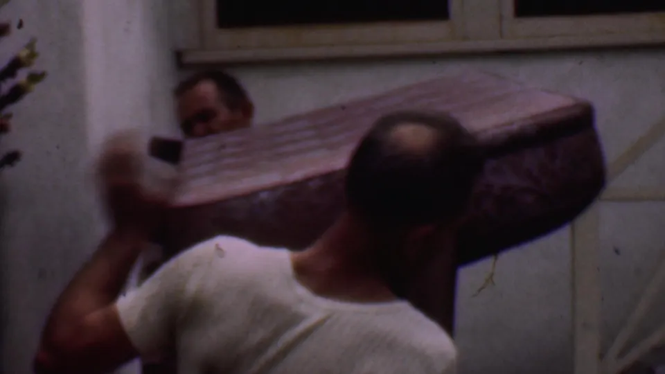 Two adult men in the Phoenix 1956 footage maneuver the patterned couch close to the camera, with the stair rail, window, and stucco wall behind them.