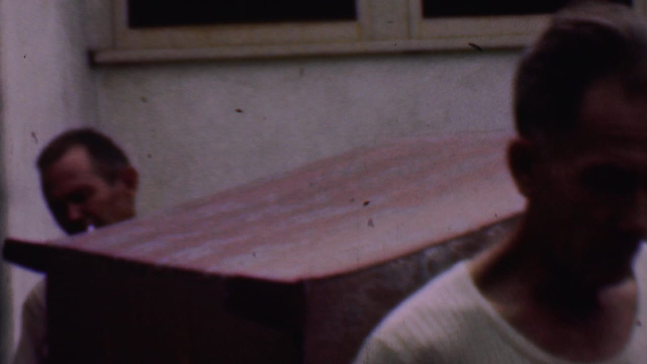 In Phoenix, Arizona home-movie footage from 1956, two adult men move a large wooden desk or cabinet past a stucco exterior wall and window.