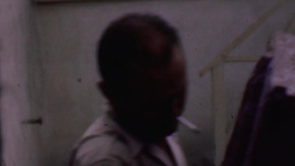 An adult man with a cigarette is seen in soft focus beside the edge of the wooden furniture during the short Phoenix moving scene.