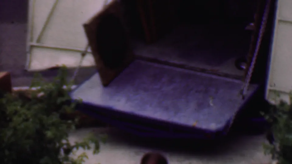 The large wooden desk or cabinet is angled near a doorway or stairwell area, with a stucco wall and small greenery visible in the final Phoenix home-movie frame.