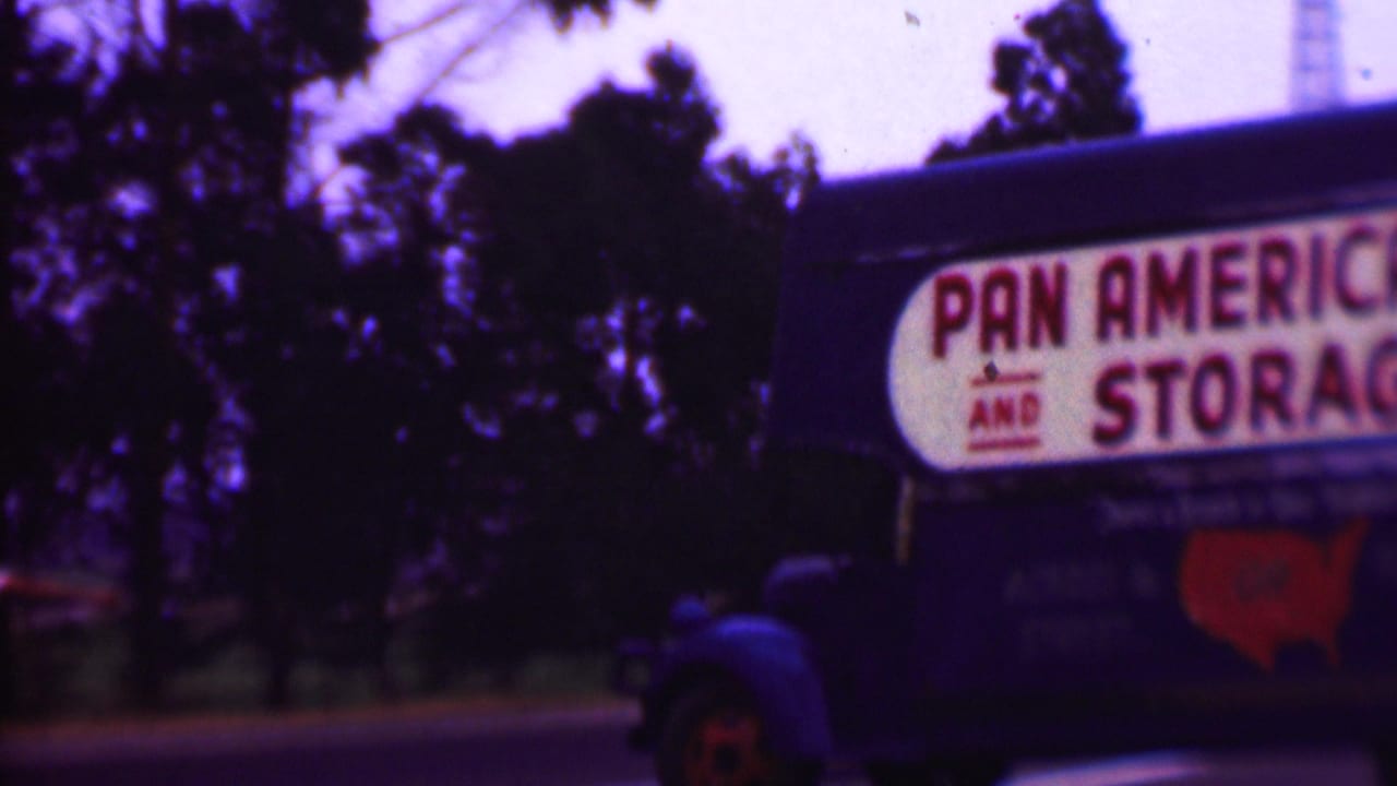 Pan American Van and Storage Co sign on the side of a blue moving truck beside roadside trees.