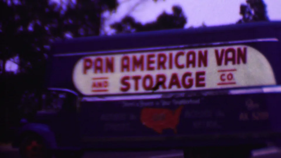 1956 Phoenix 8mm frame showing a close side view of the blue Pan American Van and Storage Co truck, with the white sign panel, red lettering, and roadside trees visible.