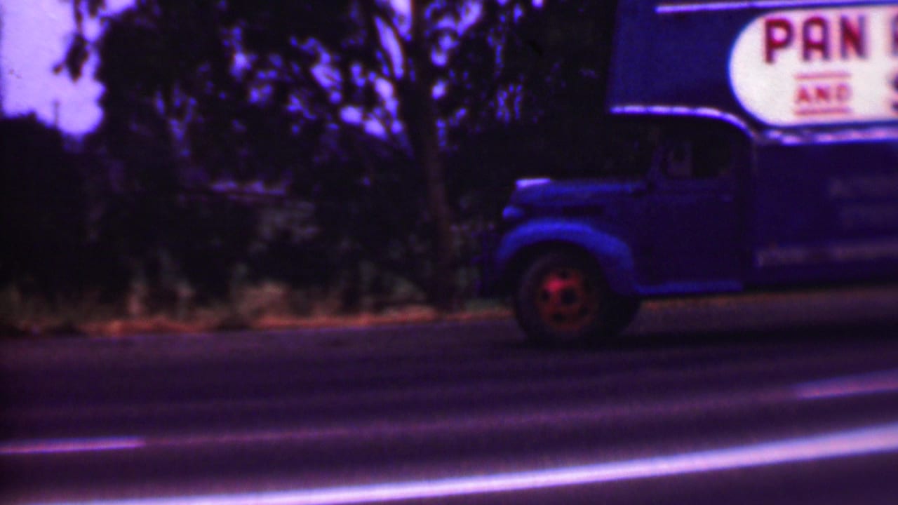 1956 Phoenix-area 8mm frame showing a blue moving truck with partial painted signage beside a road while a light-colored vintage car passes in front.