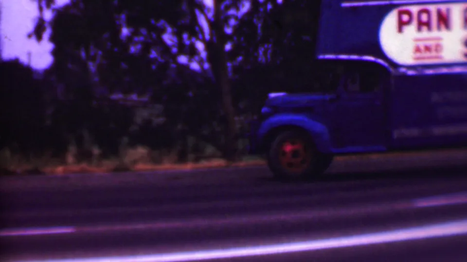 1956 Phoenix-area 8mm frame showing a blue moving truck with partial painted signage beside a road while a light-colored vintage car passes in front.