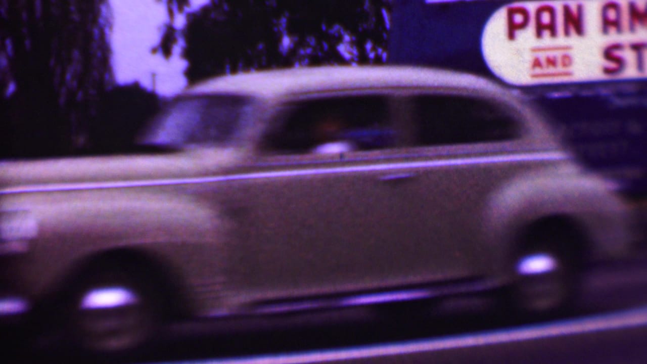 A light-colored mid-century passenger car blurs across the foreground of a Phoenix-area roadside shot with a blue moving truck and partial “PAN” signage behind it.
