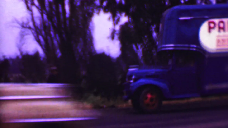 A blue moving truck stands or pulls near the roadside in a 1956 Phoenix-area home-movie frame, with trees behind and a dark blurred foreground vehicle or shadow.