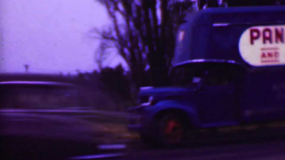 Final 1956 Phoenix-area frame showing the blue moving truck near the right side of the road, partial commercial lettering, trees, and blurred passing traffic.