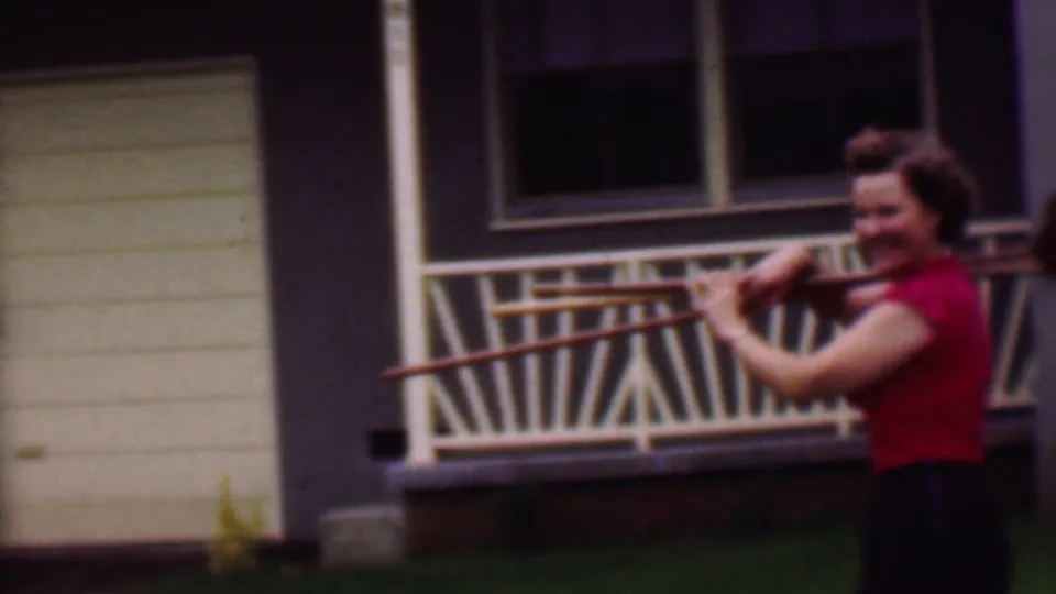 A woman in a red top walks beside a light-colored Phoenix house carrying long wooden poles, with a white garage door, railing, windows, and lawn behind her.