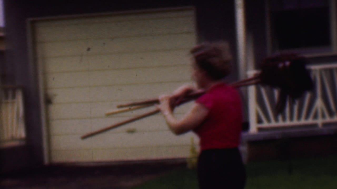 In 1956 Phoenix home-movie footage, a woman in a red shirt and dark skirt walks past a beige house and white garage door carrying mops or brooms over her shoulder.