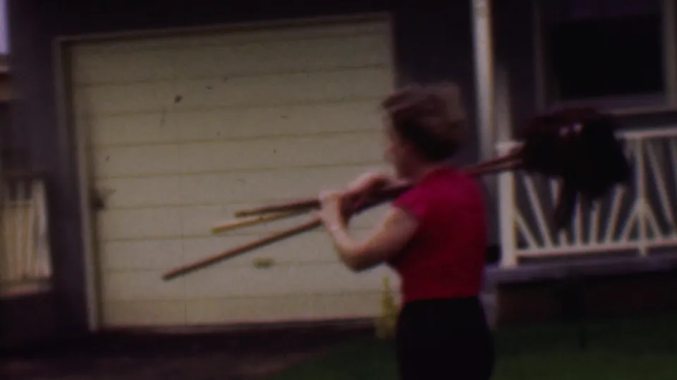 In 1956 Phoenix home-movie footage, a woman in a red shirt and dark skirt walks past a beige house and white garage door carrying mops or brooms over her shoulder.