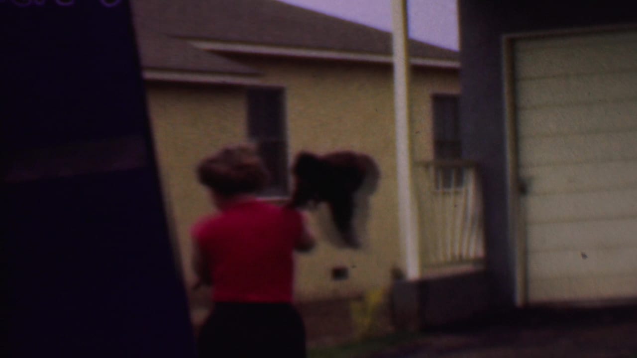 The woman continues across the residential exterior in Phoenix, carrying a bundle of mops or brooms near a dark upright structure and a beige house wall.
