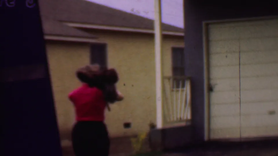 A rear view shows the woman in a red top and dark skirt walking quickly past porch railings and a beige residential wall with cleaning tools on her shoulder.