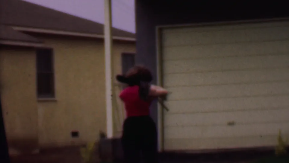 Near the end of the clip, the woman carrying mops or brooms moves beside a white garage door and house exterior in the 1956 Phoenix residential setting.