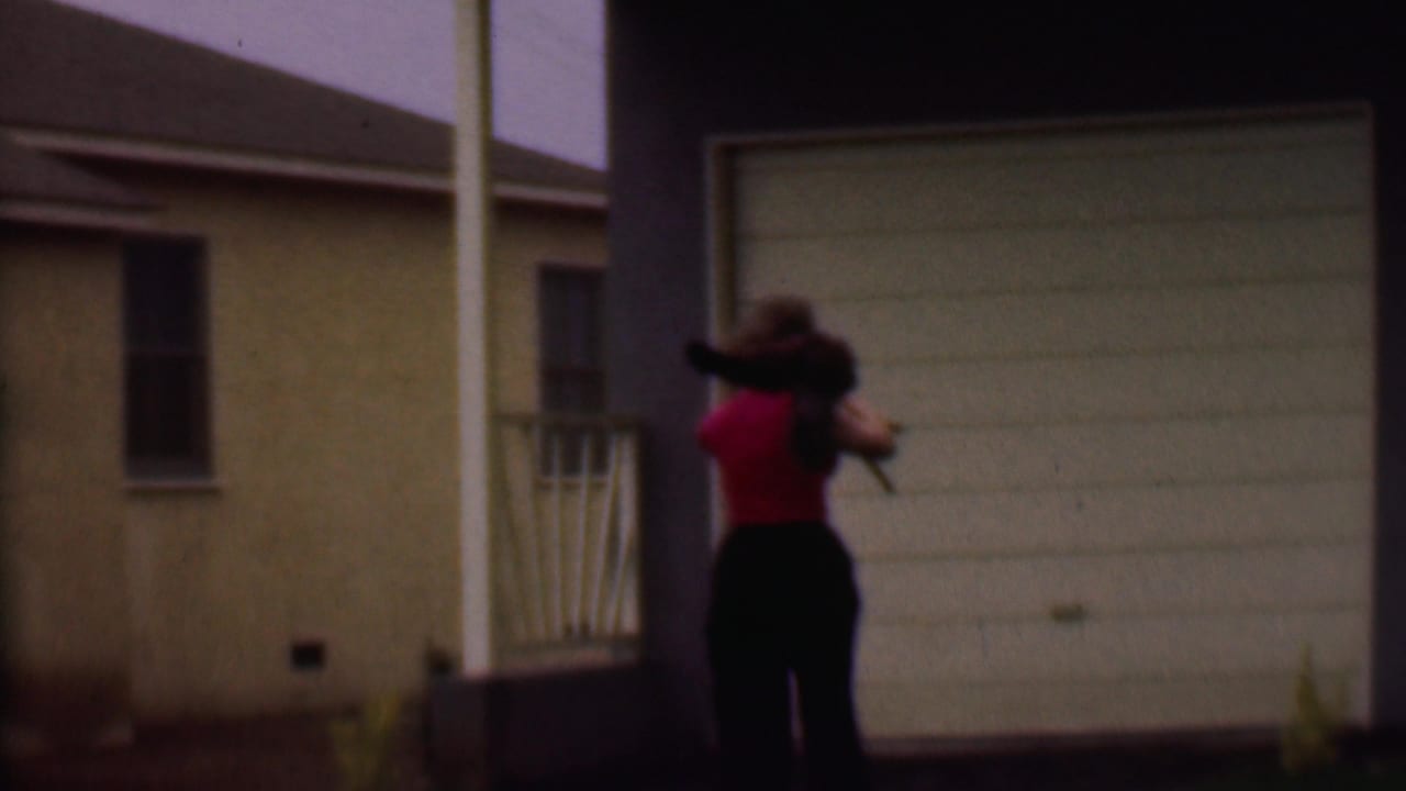 In supported 1956 Phoenix home-movie footage, a woman in a red top and dark pants carries a camera beside a suburban house with a beige garage door.