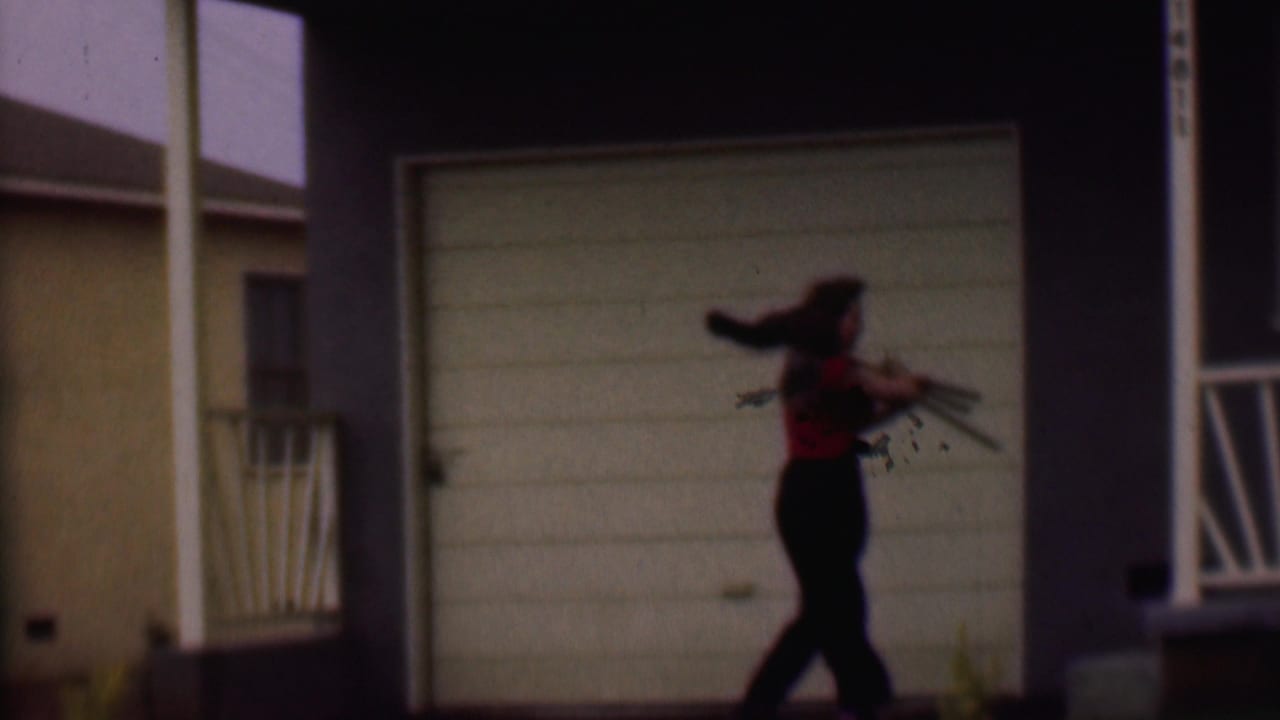 The woman continues walking across the suburban house frontage in Phoenix, carrying a camera near the garage and porch area.