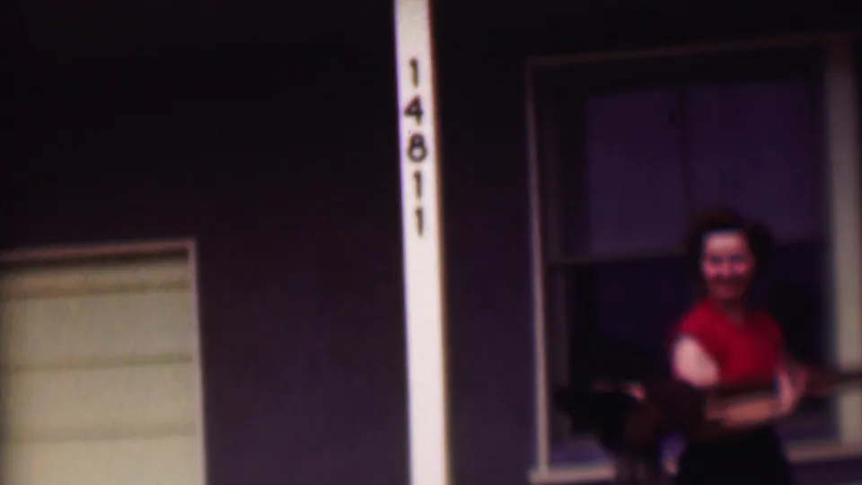 A smiling woman with a camera approaches the porch area of the Phoenix house, with the number 14811 visible on a vertical post beside her.