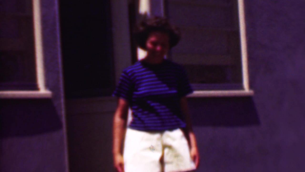 In supported 1956 Phoenix home-movie footage, a woman in a blue striped shirt and white shorts stands on concrete steps near a doorway and window in bright sunlight.