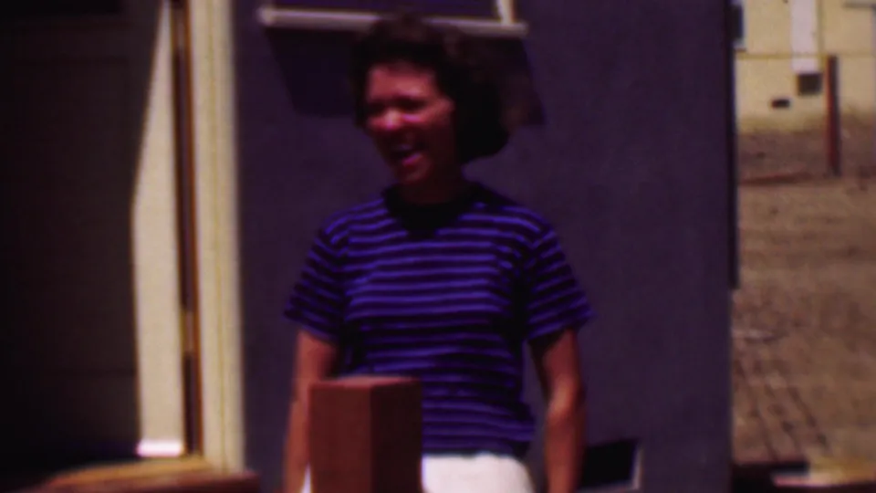 A smiling woman in a blue striped top is framed closer outside the building, with the door edge, stucco-like wall, and sunlit ground behind her.