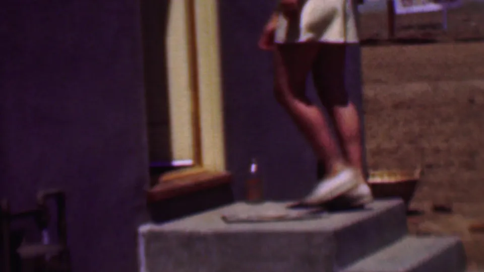 The lower half of the woman is visible as she steps down from a small concrete stoop, showing white shorts, bare legs, white shoes, the doorway, and dry ground nearby.