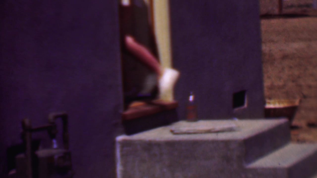 Phoenix 1956 8mm frame showing a cropped person in white shoes on a concrete step beside a doorway and exterior wall.