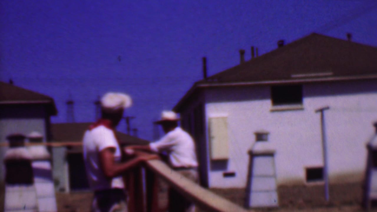 Phoenix 1956 8mm frame showing two adult men in white caps working or standing by a wooden railing in front of white residential buildings.
