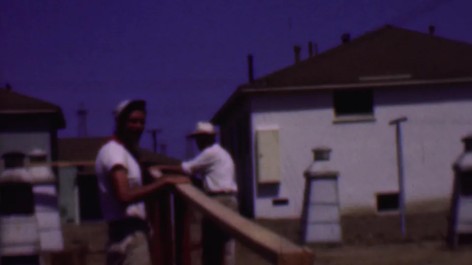 Phoenix 1956 8mm frame showing one man near a wooden railing looking toward the camera, with another man farther back near the white house exterior.