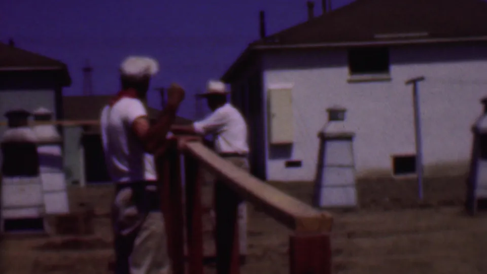 Phoenix 1956 8mm frame showing two adult men in white caps beside a long wooden railing, with white residential buildings and dry ground behind them.