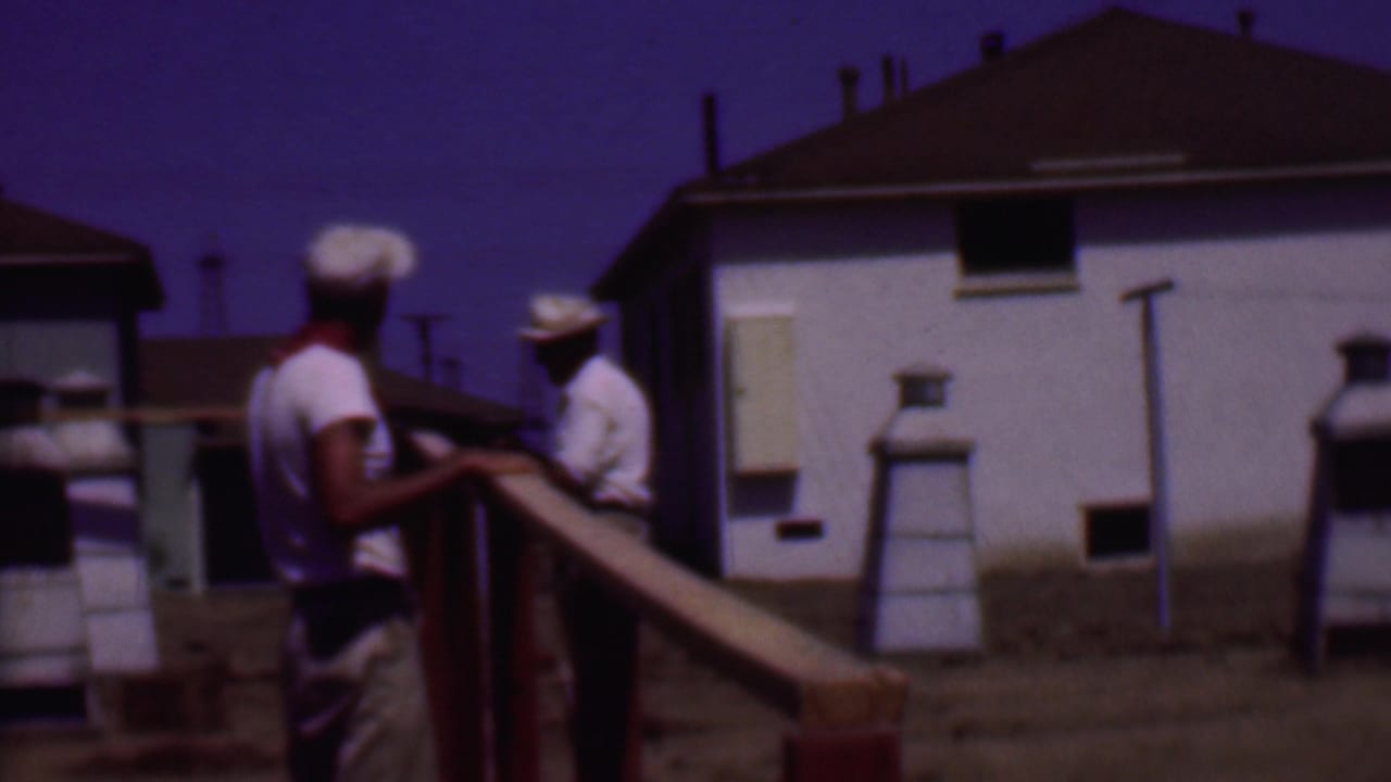 Phoenix, Arizona 1956 home-movie frame showing two men in light shirts and hats working beside a wooden wall frame in a sunlit residential area.