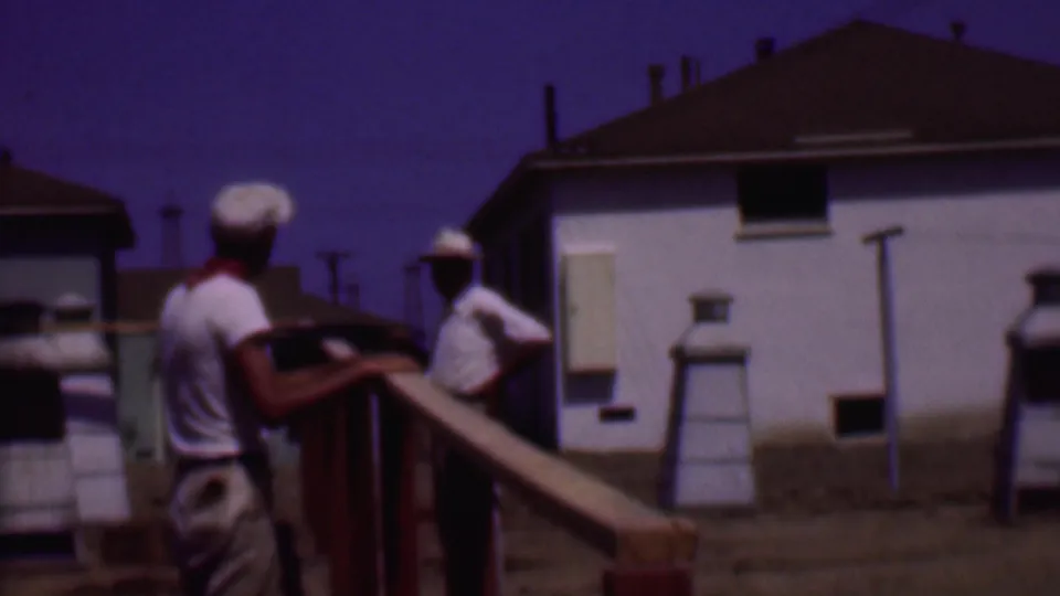 Two men in hats continue work on a wooden frame in a sunlit Phoenix residential setting, with houses and yard structures visible behind them.