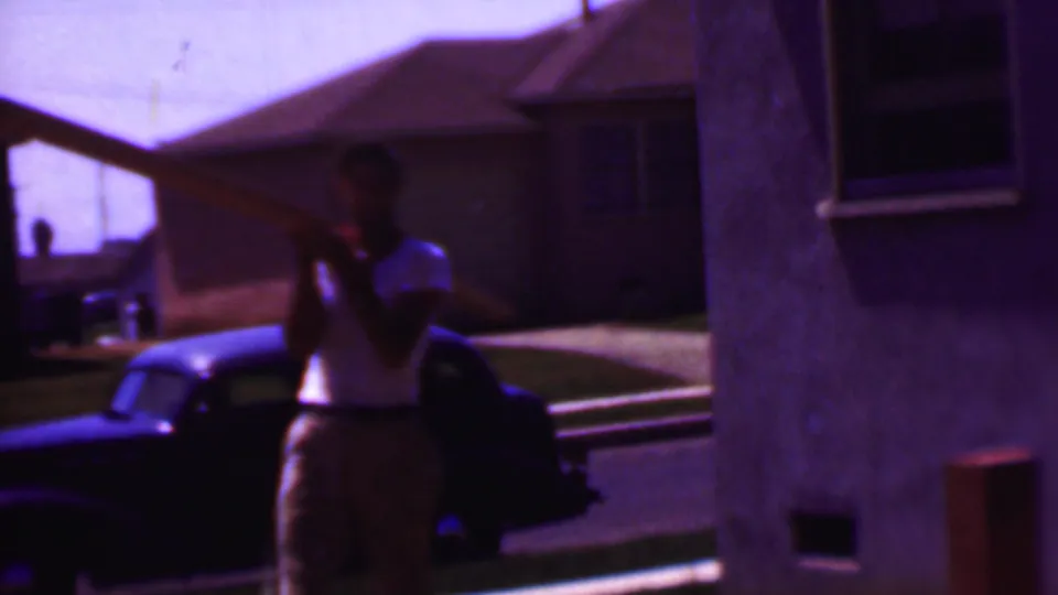 A man carries a large wooden panel or frame near a parked dark car and residential houses in the 1956 Phoenix home-movie footage.