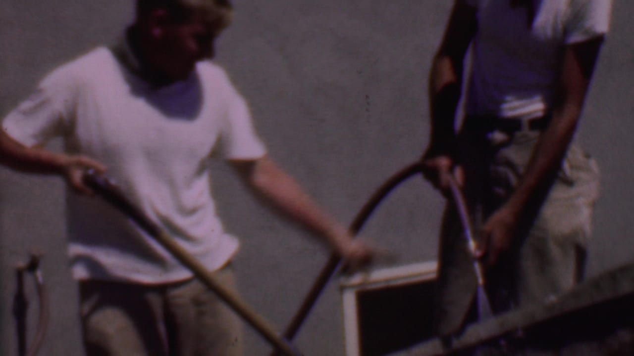 In 1956 Phoenix home-movie footage, two adult men stand outdoors by a wheelbarrow as one mixes cement with a shovel and the other handles a hose.