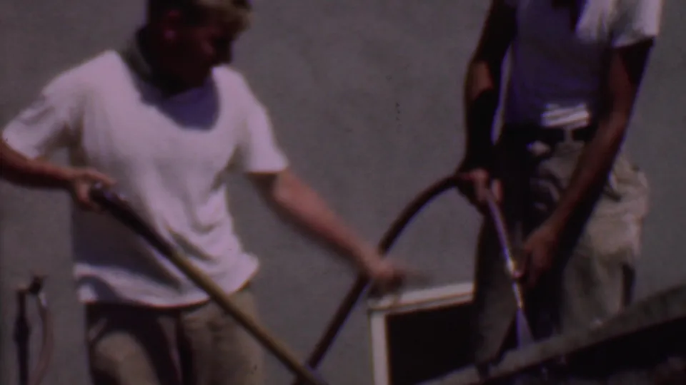 In 1956 Phoenix home-movie footage, two adult men stand outdoors by a wheelbarrow as one mixes cement with a shovel and the other handles a hose.