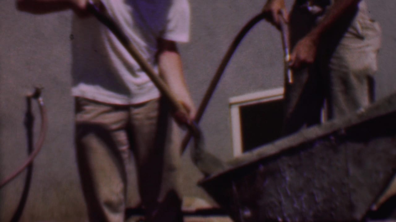 A man holds a hose over a wheelbarrow while another man stands nearby with a shovel.