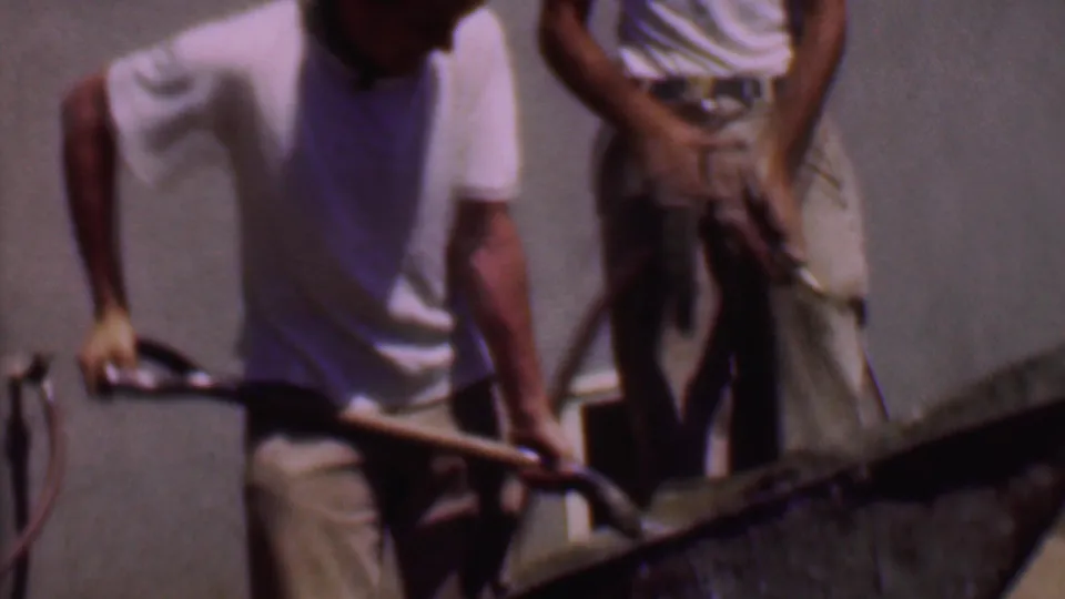 The 8mm frame shows a man leaning into the work of mixing cement in a wheelbarrow while another adult man stands close by against a plain wall.