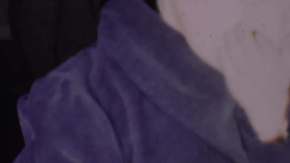 A close 1956 Phoenix home-movie frame shows part of an adult woman's pale facial profile beside a bulky blue-purple draped covering against a dark interior background.