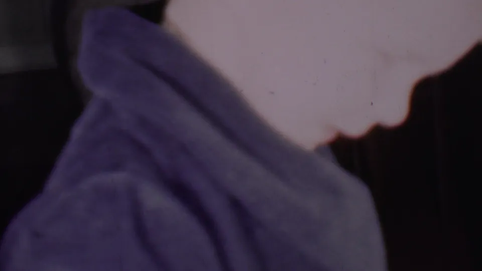 A close 1956 Phoenix home-movie frame shows an adult woman leaning downward in partial profile, with deep blue-purple fabric wrapped around her head and shoulders.