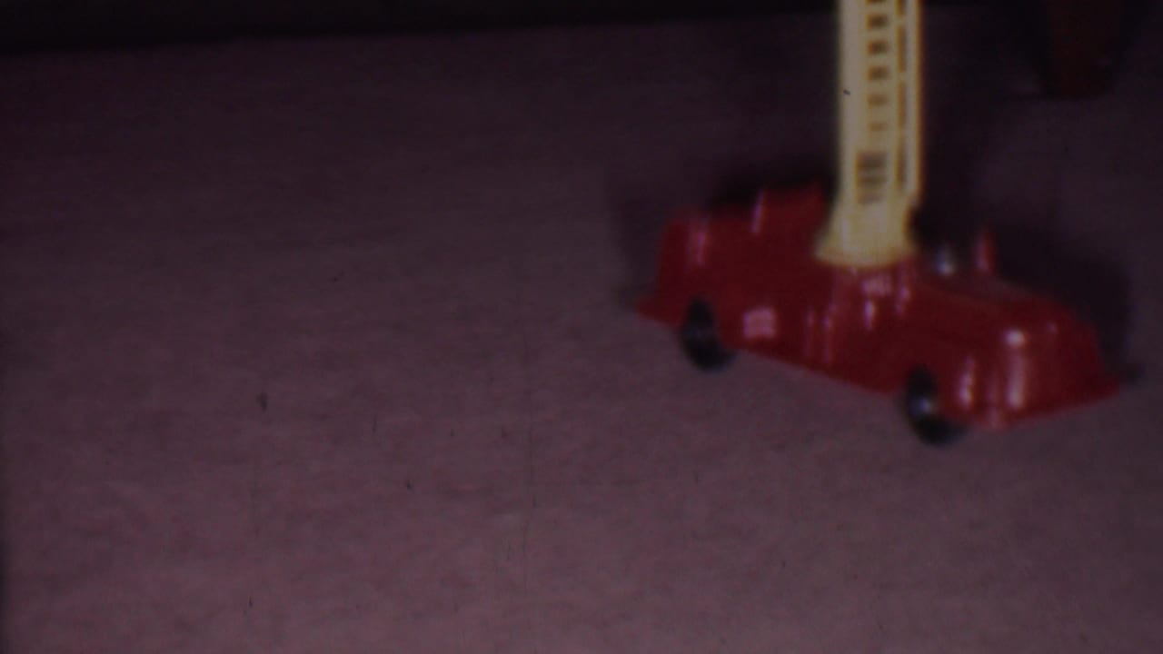 A red toy fire engine with a yellow ladder sits on carpet in the Phoenix 1956 home-movie scene, with the frame softly exposed and partially cropped.
