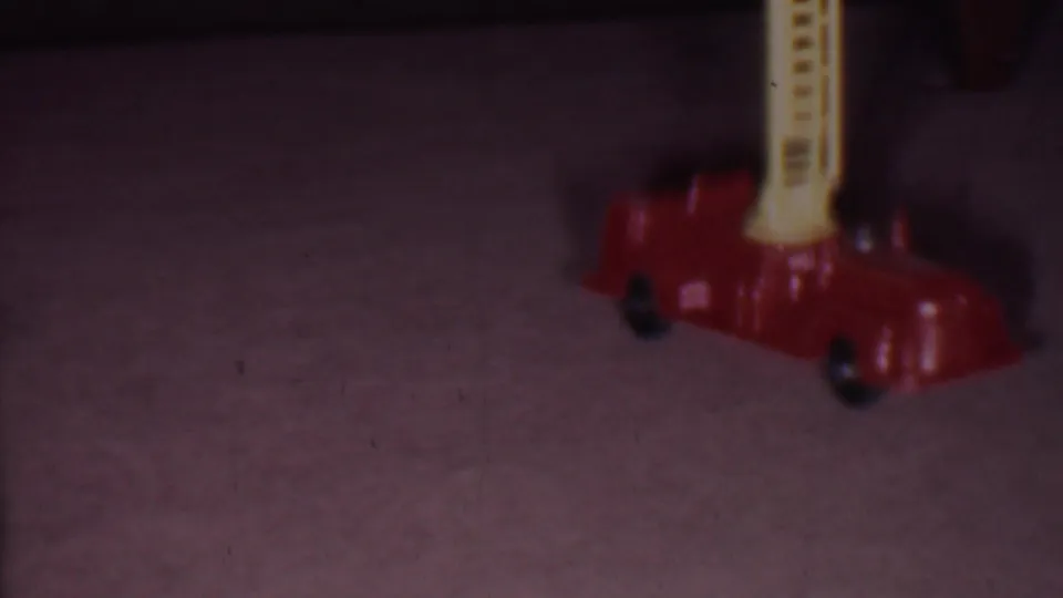 A red toy fire engine with a yellow ladder sits on carpet in the Phoenix 1956 home-movie scene, with the frame softly exposed and partially cropped.