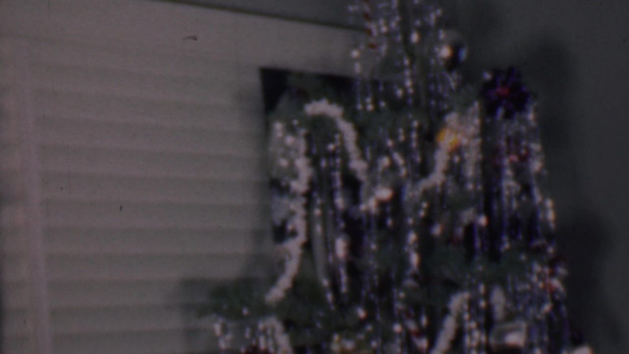 Phoenix, Arizona 1956 home-movie frame showing the upper part of an indoor Christmas tree with white garland, metallic tinsel, dark ornaments, and horizontal blinds in the background.