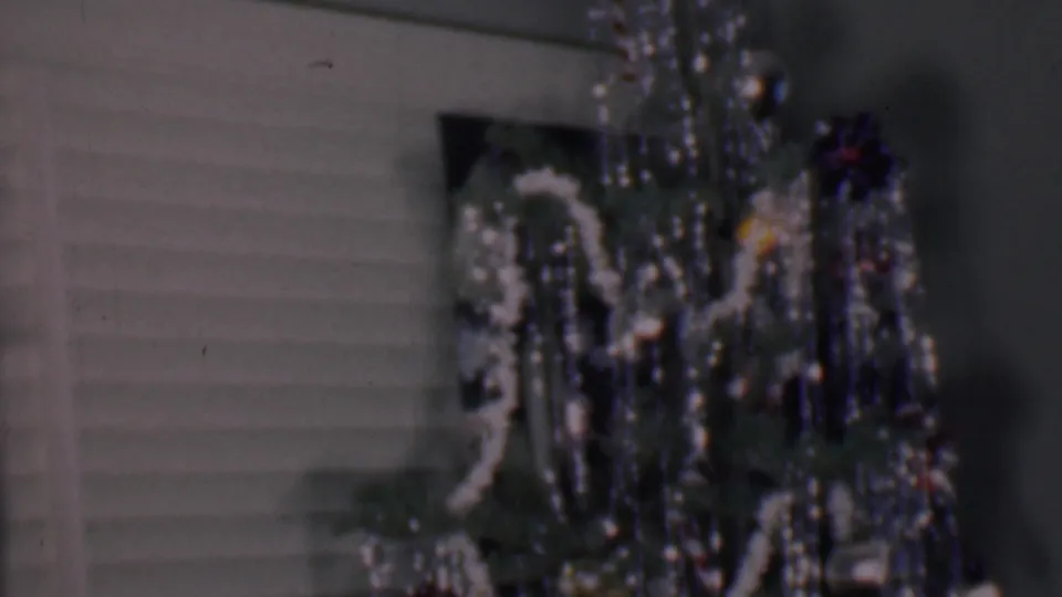 Phoenix, Arizona 1956 home-movie frame showing the upper part of an indoor Christmas tree with white garland, metallic tinsel, dark ornaments, and horizontal blinds in the background.