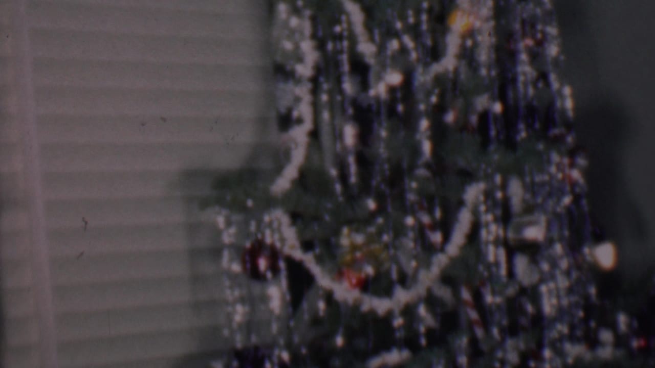 Decorated Christmas tree section with white garland, tinsel, ornaments, and pale blinds behind it.