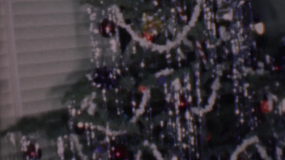 Phoenix, Arizona 1956 frame with the camera close to a tinsel-covered indoor Christmas tree, showing white garland, red ornaments, small lights, and horizontal blinds.