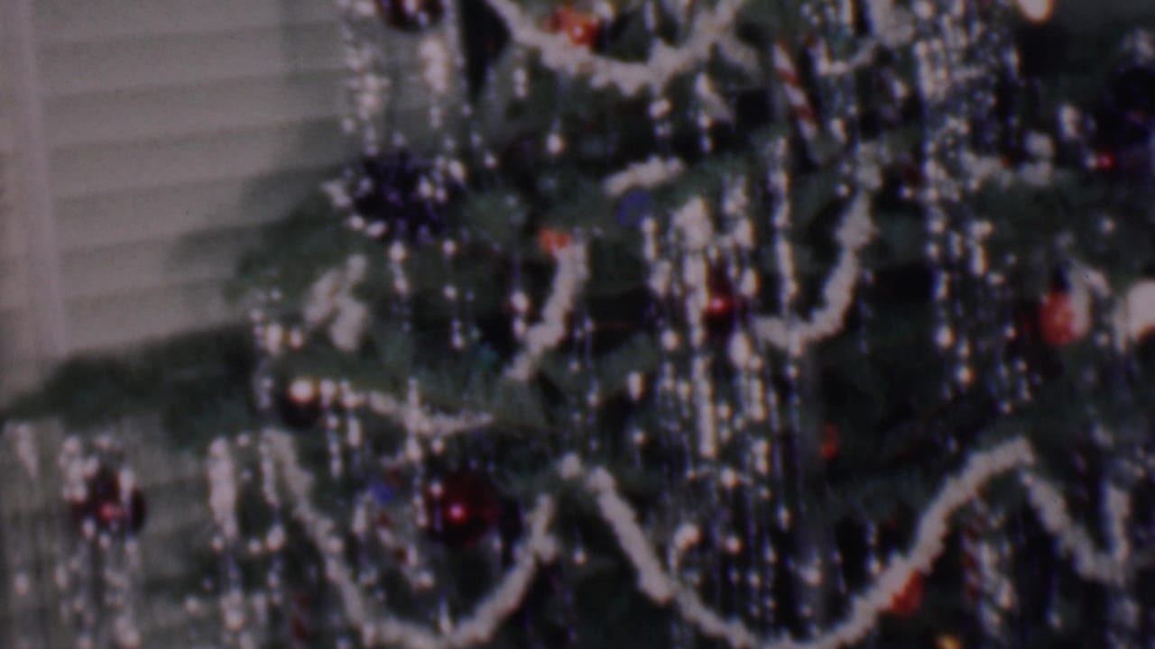 Close 1956 Phoenix home-movie view of a decorated Christmas tree with white garland, red ornaments, reflective strands, and horizontal blinds behind it.