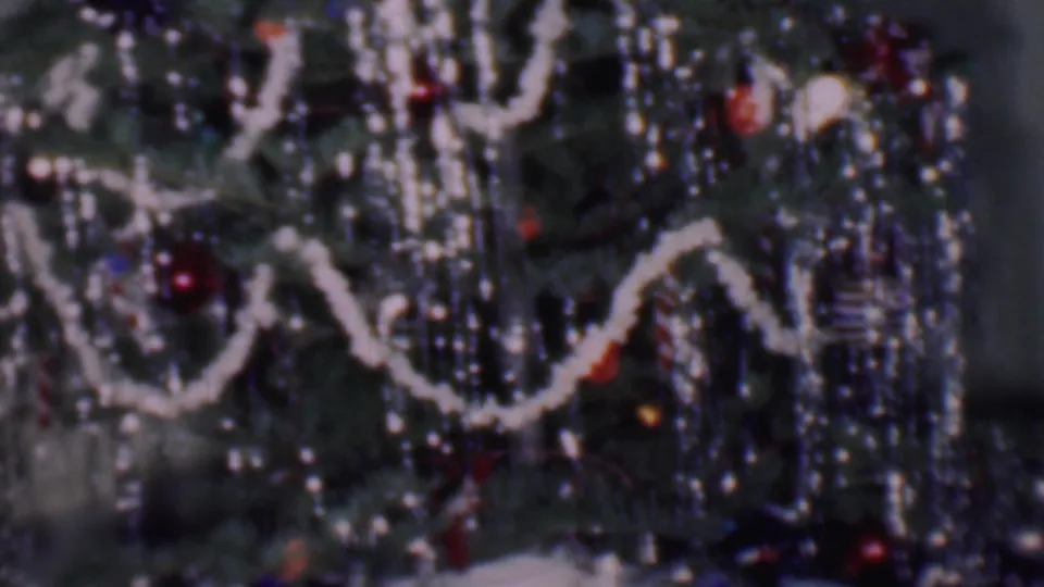Close 1956 Phoenix home-movie frame showing a decorated Christmas tree with white garland, reflective hanging strands, ornaments, horizontal blinds, and a pale tree skirt near the bottom.