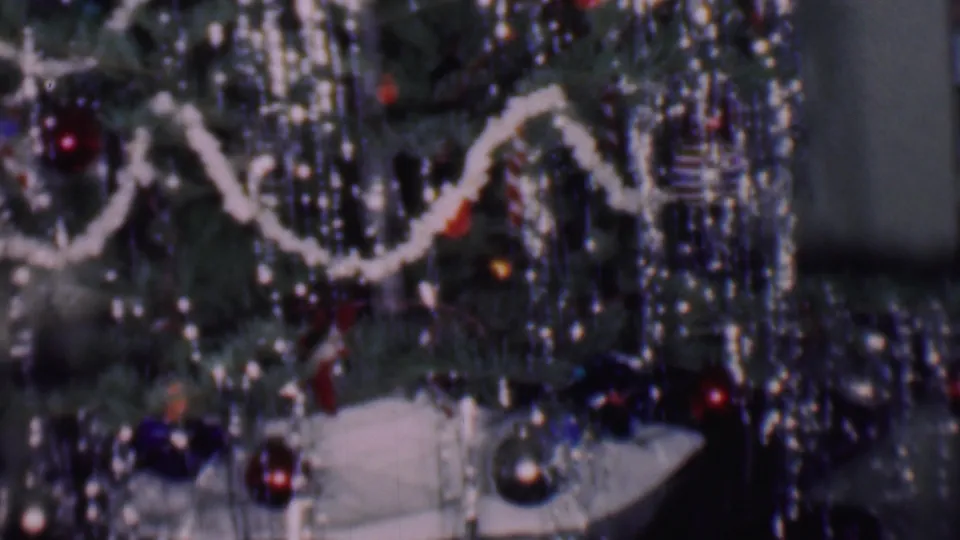 Close 1956 Phoenix home-movie frame of a decorated Christmas tree with white garland, reflective strands, red and dark ornaments, and a white tree skirt below.