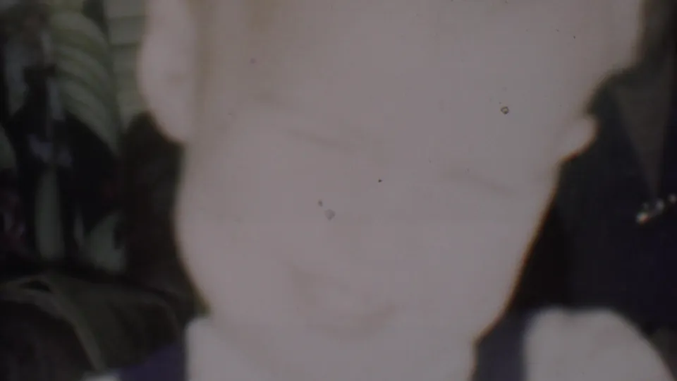 Faded 8mm home-movie frame showing a pale, soft-focus close-up shape beside dark foliage or decoration, with visible dust and exposure variation.