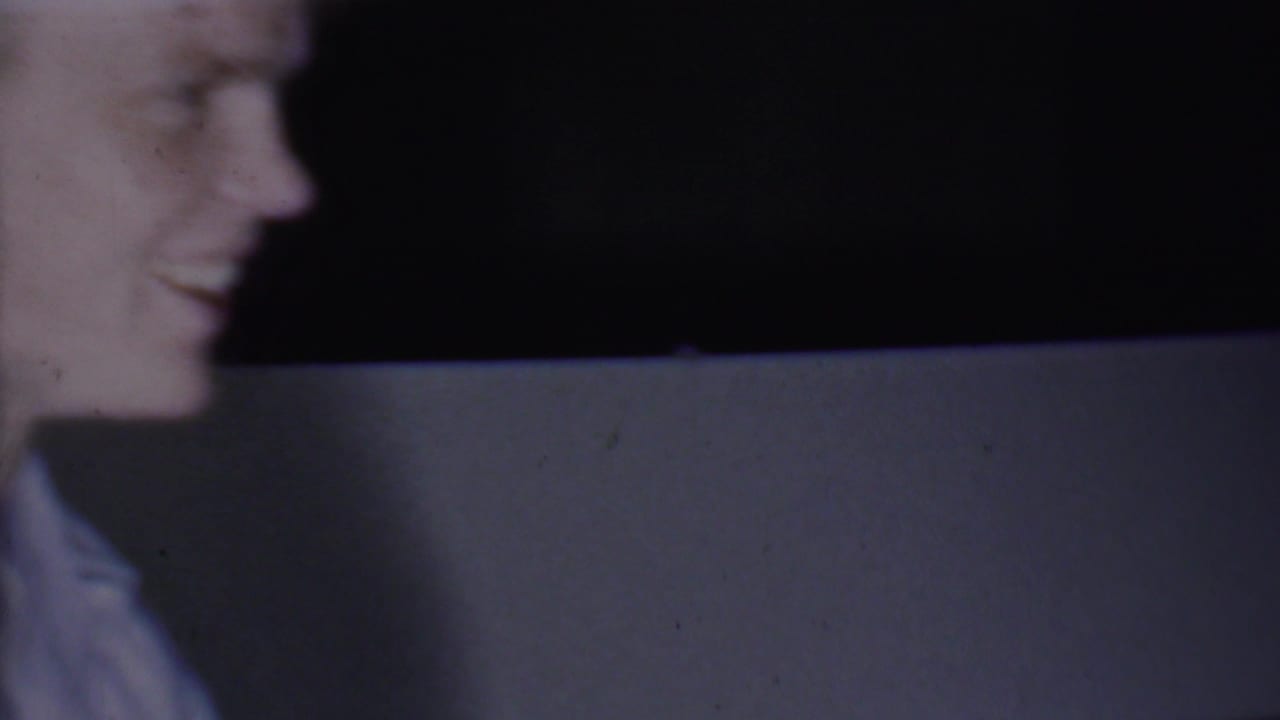 In this 1956 Phoenix home-movie frame, a smiling adult man is partly visible at the left edge against a pale wall and dark background.