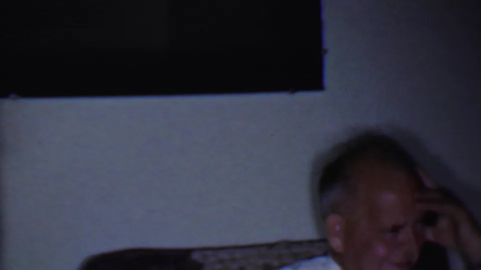 In this Phoenix, Arizona 1956 frame, a seated adult man laughs in a low-lit private room with wall surfaces and furniture partly visible.