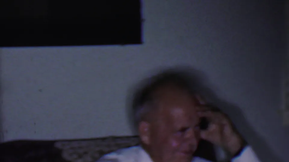 The 1956 Phoenix footage shows a blurred adult man laughing in a private interior, with pale wall space and furniture behind him.